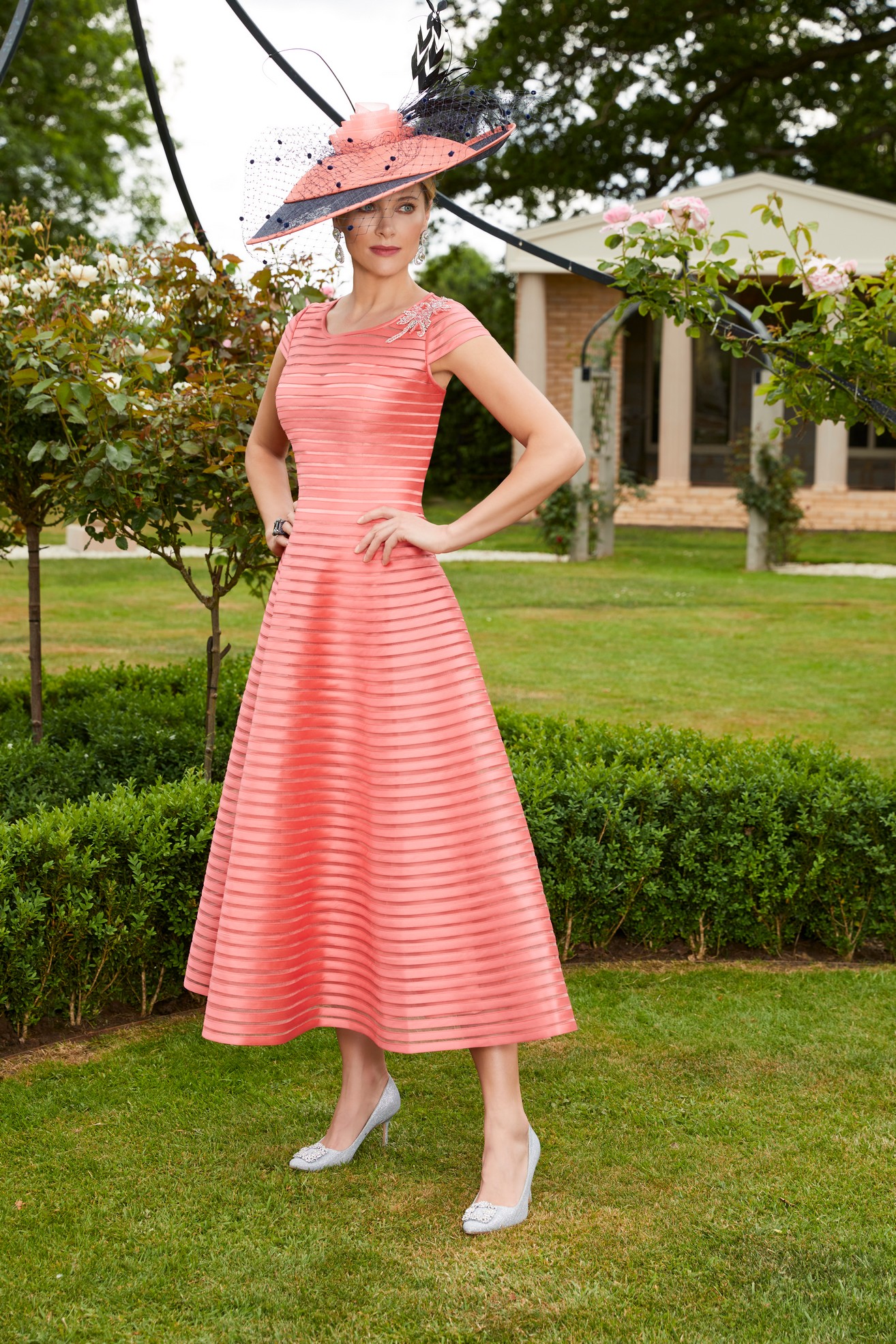 Lady in coral pink tea style a-line dress with cap sleeves and round neckline with matching fascinator hat and high heeled shoes standing in garden 